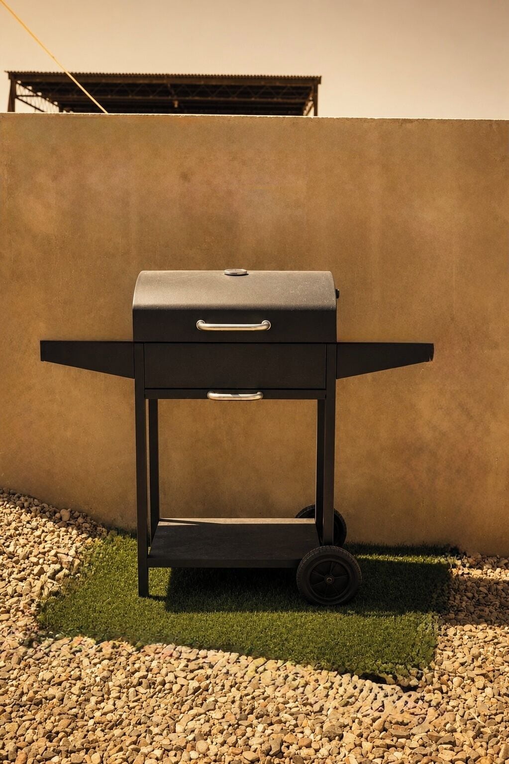 A black barbecue grill on a wheeled cart, positioned on a gravel surface with grass, against a plain wall.