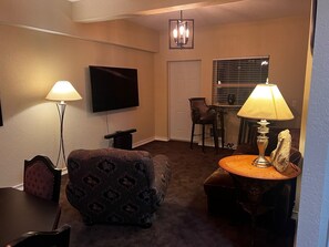 Ein Wohnzimmer mit Flachbildfernseher, einem gemusterten Sessel, einem kleinen Tisch mit Lampe und einem Essbereich mit Fenster.