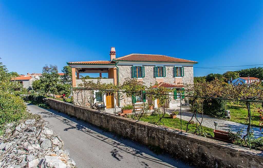 A two-story house with a tiled roof, a stone wall in front, and a garden with potted plants.