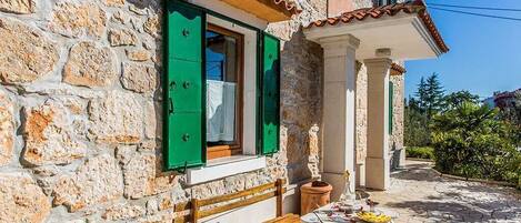 A stone building with green shutters, a wooden bench, and a table with a plate of food.