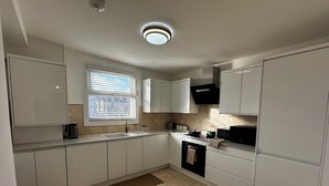 A modern kitchen with white cabinets, a black microwave, and a window with blinds. - Charming 4-bedroom apartment in lively London neighborhood (London)