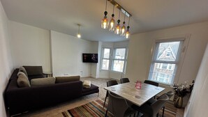 A living room with a sofa, a dining table with chairs, a TV mounted on the wall, and large windows with blinds. - Charming 4-bedroom apartment in lively London neighborhood (London)