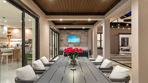A modern dining area with a long table, white cushioned chairs, and a vase of red flowers. - 1-Acre Oasis: Pool/Spa, Pickleball, Bball & More (Miami)