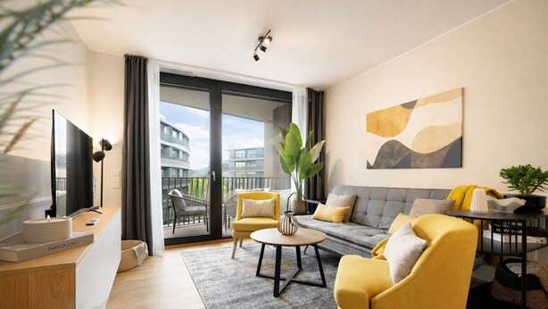 A modern living room with a sofa, armchairs, a coffee table, a TV, and a balcony with a view of buildings. - Gleam Homes Marina Garden Dresden (Dresden)