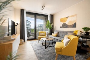 A modern living room with a sofa, armchairs, a coffee table, a TV, and a balcony with a view of buildings. - Gleam Homes Marina Garden Dresden (Dresden)