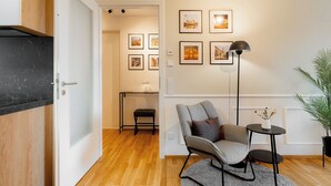 A modern living room with a wooden floor, a grey armchair, a small round table with a lamp, and framed pictures on the wall. - Gleam Homes Marina Garden Dresden (Dresden)