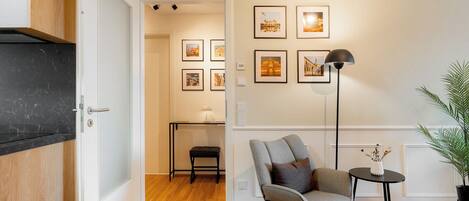 A modern living room with a wooden floor, a grey armchair, a small round table with a lamp, and framed pictures on the wall.