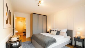 A modern bedroom with a large bed, bedside tables, a wardrobe, and a hallway leading to a bathroom. - Gleam Homes Marina Garden Dresden (Dresden)