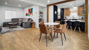 Een moderne woonkamer met een grijze bank, een ronde salontafel, en een eethoek met een witte tafel en vier stoelen.