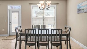 A dining area with a table and chairs, a chandelier, a door, and a window with blinds. - Beach Haven: 6 Beds, Pet Friendly, 8miles to Beach (Mary Esther)