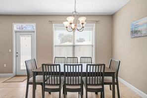 A dining area with a table and chairs, a chandelier, a door, and a window with blinds. - Beach Haven: 6 Beds, Pet Friendly, 8miles to Beach (Mary Esther)