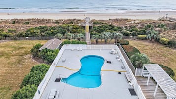 Ein Schwimmbecken mit Holzterrasse, umgeben von einem Zaun und Palmen, mit Blick auf einen Strand.