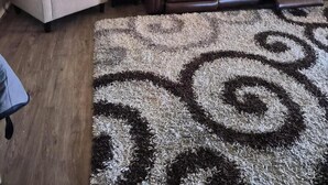 Un salon avec un fauteuil beige, un canapé en cuir marron, un tapis à motifs, un ventilateur de plafond et un tableau représentant un paysage urbain accroché au mur.