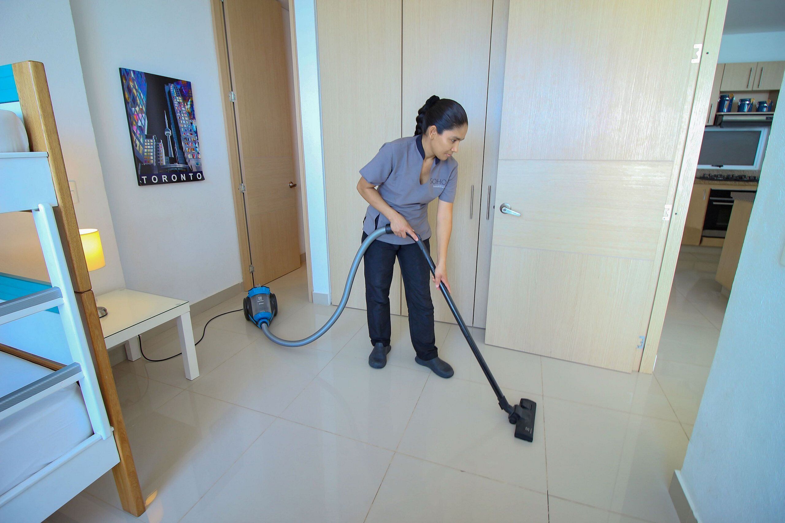 Una persona aspirando una habitación con una cama, una mesita y un cuadro colgado en la pared.