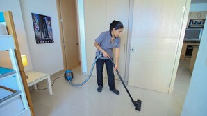 Una persona aspirando una habitación con una cama, una mesita y un cuadro colgado en la pared.