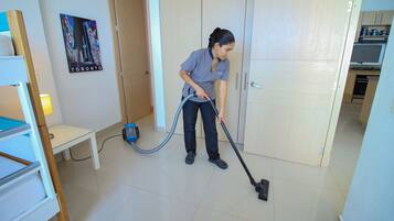 Una persona aspirando una habitación con una cama, una mesita y un cuadro colgado en la pared.