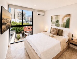 A bedroom with a bed, a nightstand, a wall-mounted air conditioner, a window with a city view, and a framed abstract painting.