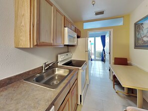 A compact kitchen with wooden cabinets, a stainless steel sink, a microwave, and a stove. There is a dining area with a table and chairs. - Harbour Beach Resort 105 (Daytona Beach)