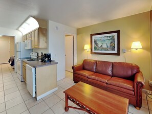 A compact living space with a kitchenette, a brown leather sofa, a wooden coffee table, and two wall-mounted lamps. - Harbour Beach Resort 205 (Daytona Beach)