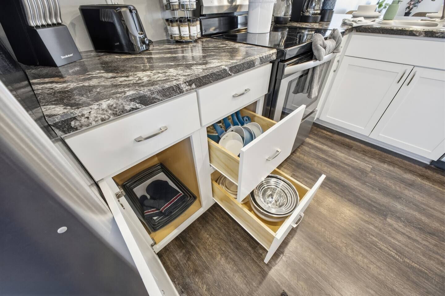 Una cocina con armarios blancos, encimera de granito y un cajón con platos y utensilios.