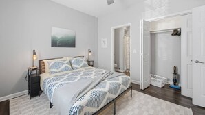 Quarto com cama, mesas de cabeceira, ventilador de teto, um quadro e um banheiro visível através de uma porta aberta.