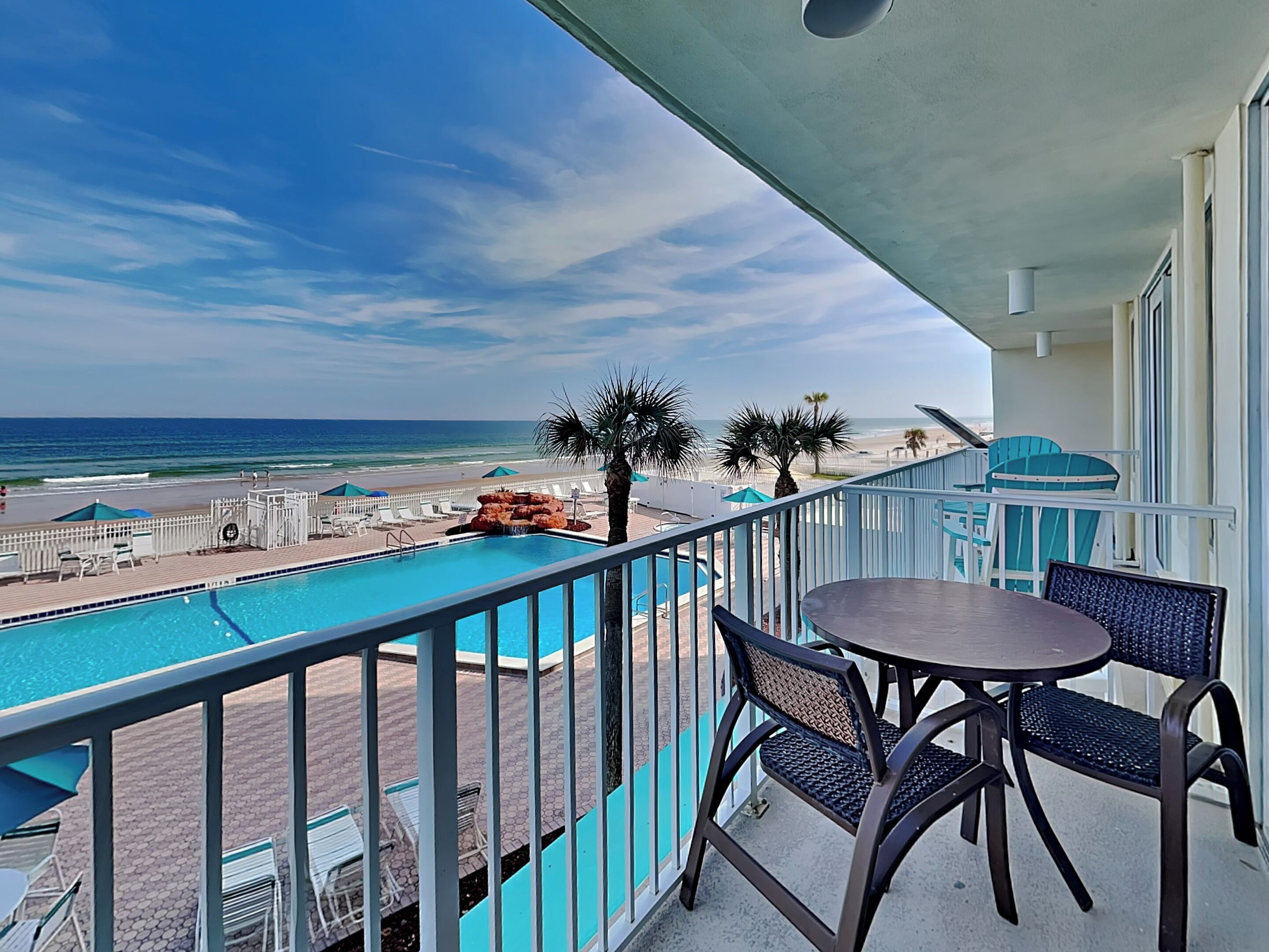 A balcony with a view of the ocean, a pool, and outdoor seating.