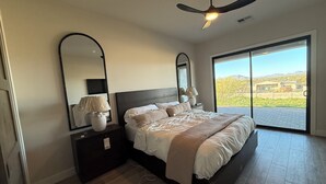 Ein Schlafzimmer mit einem großen Bett, einem Deckenventilator, einem Nachttisch mit Lampe, einem Spiegel und Blick auf eine Landschaft durch eine Schiebetür.
