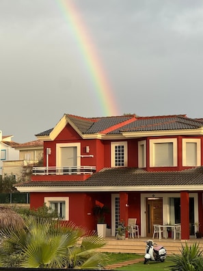 A two-story house with a red exterior, a tiled roof, and a visible rainbow in the background. - Red House Rooms  (Llucmajor)