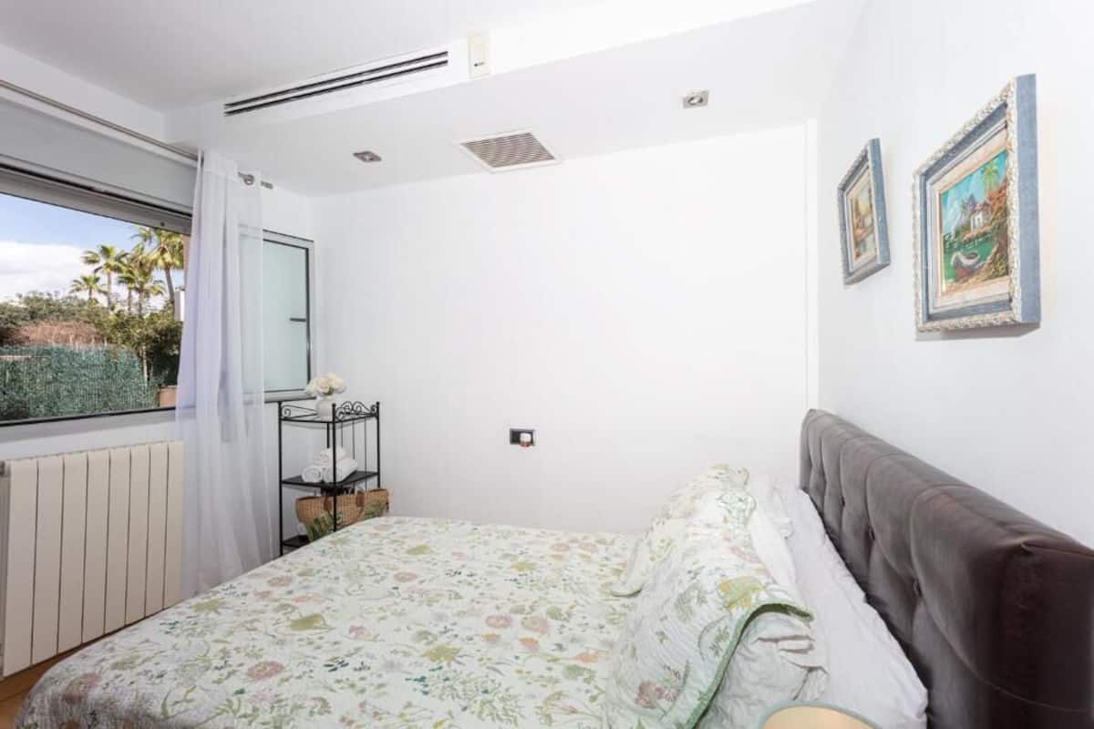 A bedroom with a bed, a window with a view of palm trees, a radiator, and a shelf with decorative items.