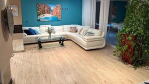 A modern living room with a white sectional sofa, a glass coffee table, and a large painting of a coastal landscape on the teal wall. - Red House Rooms  (Llucmajor)
