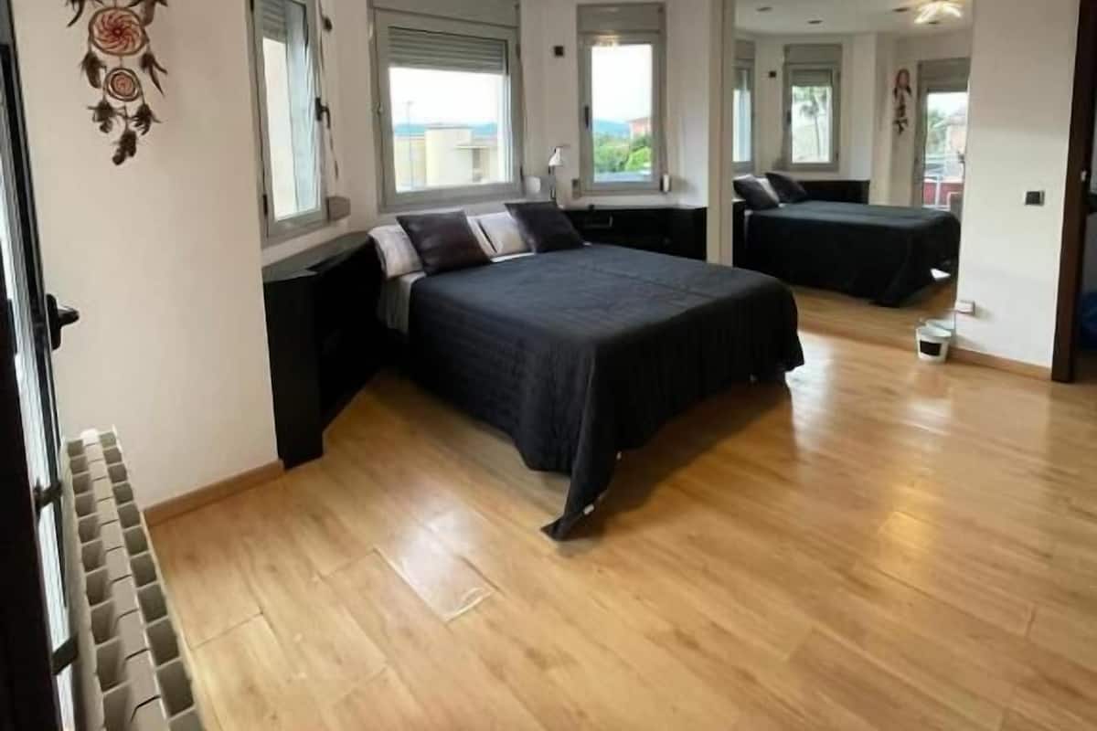 A spacious bedroom with a large bed, a desk, and a chair. There are two windows providing natural light and a view of the outside.