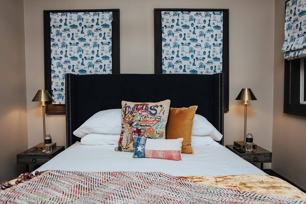 Een net opgemaakt bed met witte lakens, een kleurrijk kussen en een gebogen deken. Twee nachtkastjes met lampen en een waterfles staan erbij. De kamer heeft een donker hoofdbord en kunstwerken boven het bed.
