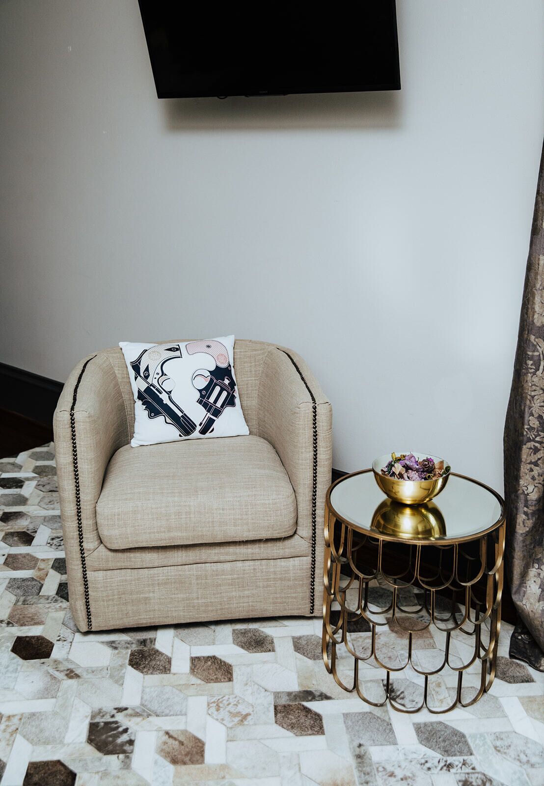 A beige sofa with a decorative pillow, a round gold side table with a bowl of flowers, and a tiled floor.