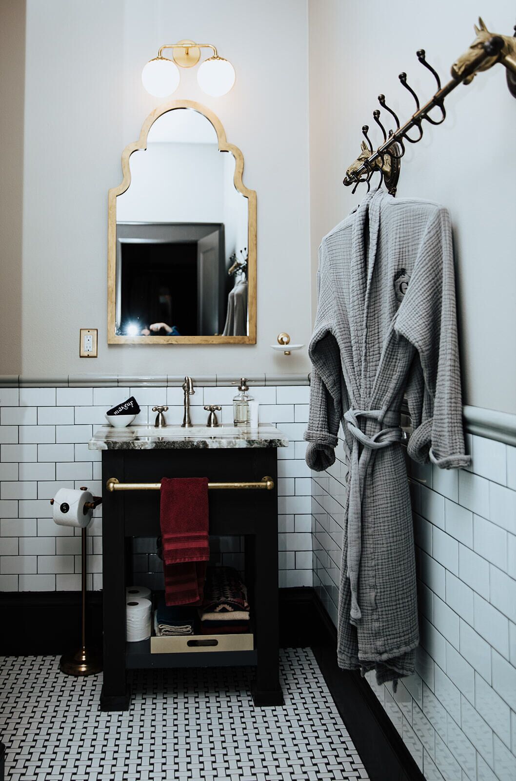 Ein Badezimmer mit Waschtisch, einem Spiegel mit goldener Rahmung, einem Handtuchhalter mit Bademantel und einem gemusterten Boden.