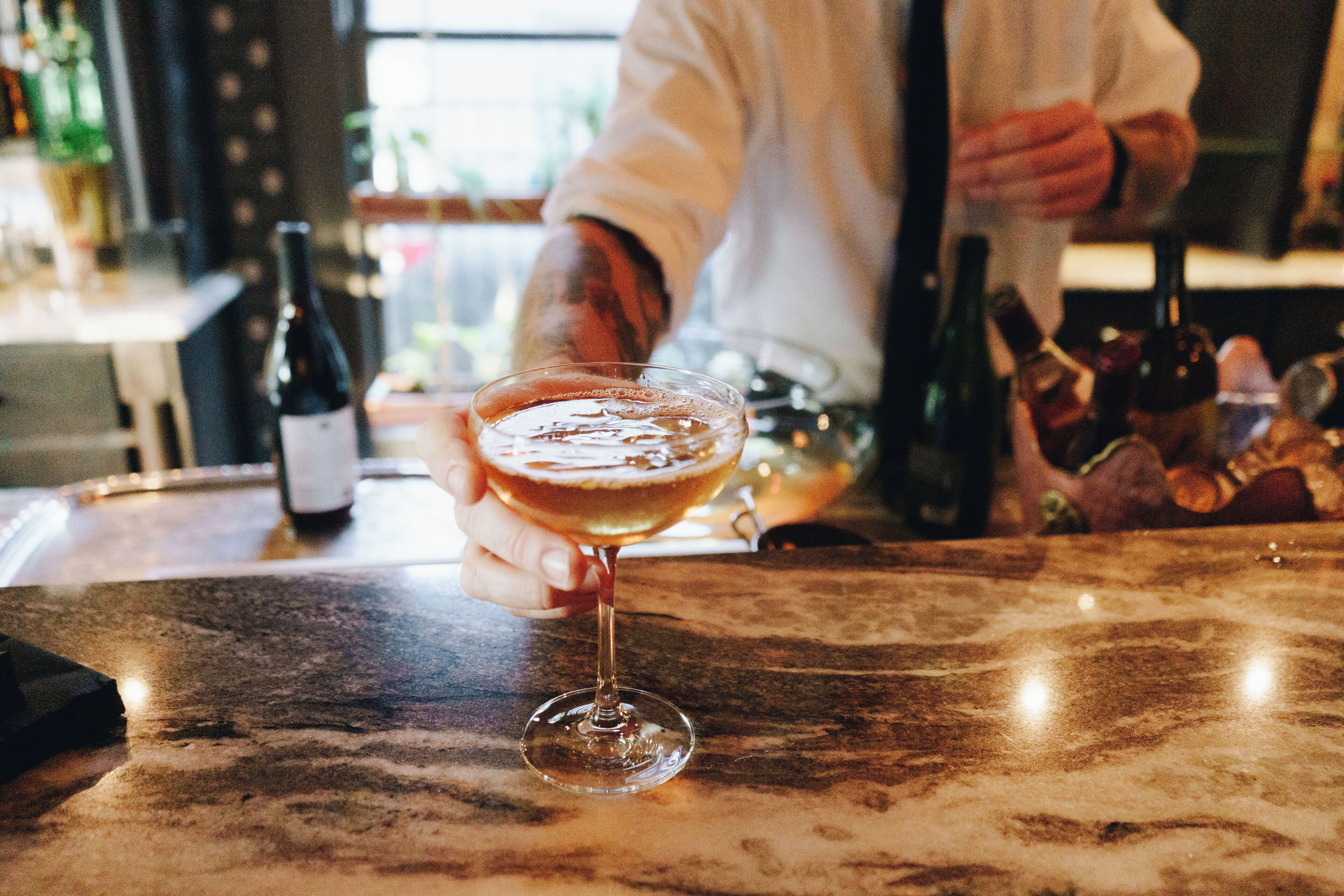 A bar setting with a glass of a light-colored beverage, a bottle of wine, and a person in the background.