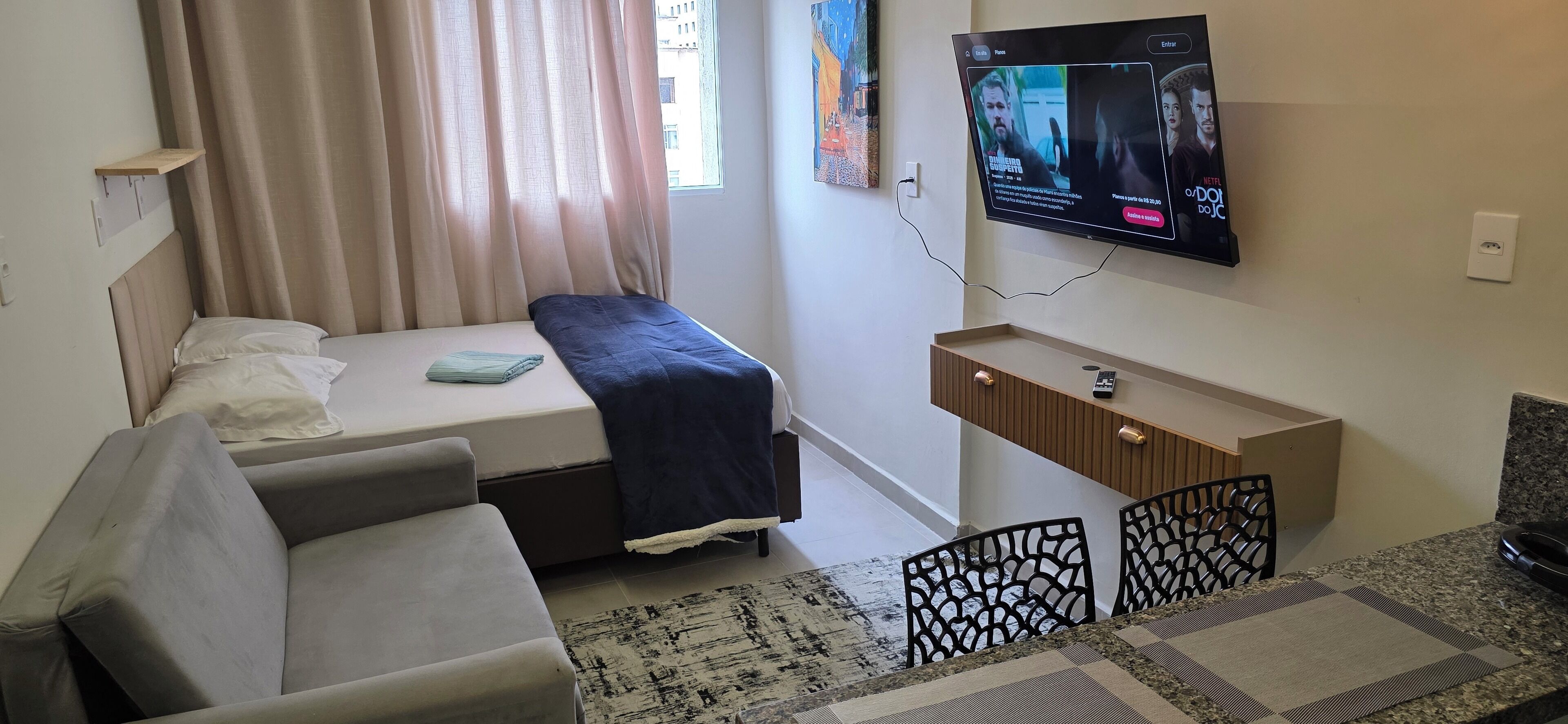 A hotel room with a bed, a sofa, a TV mounted on the wall, a small wooden cabinet, and a window with curtains.