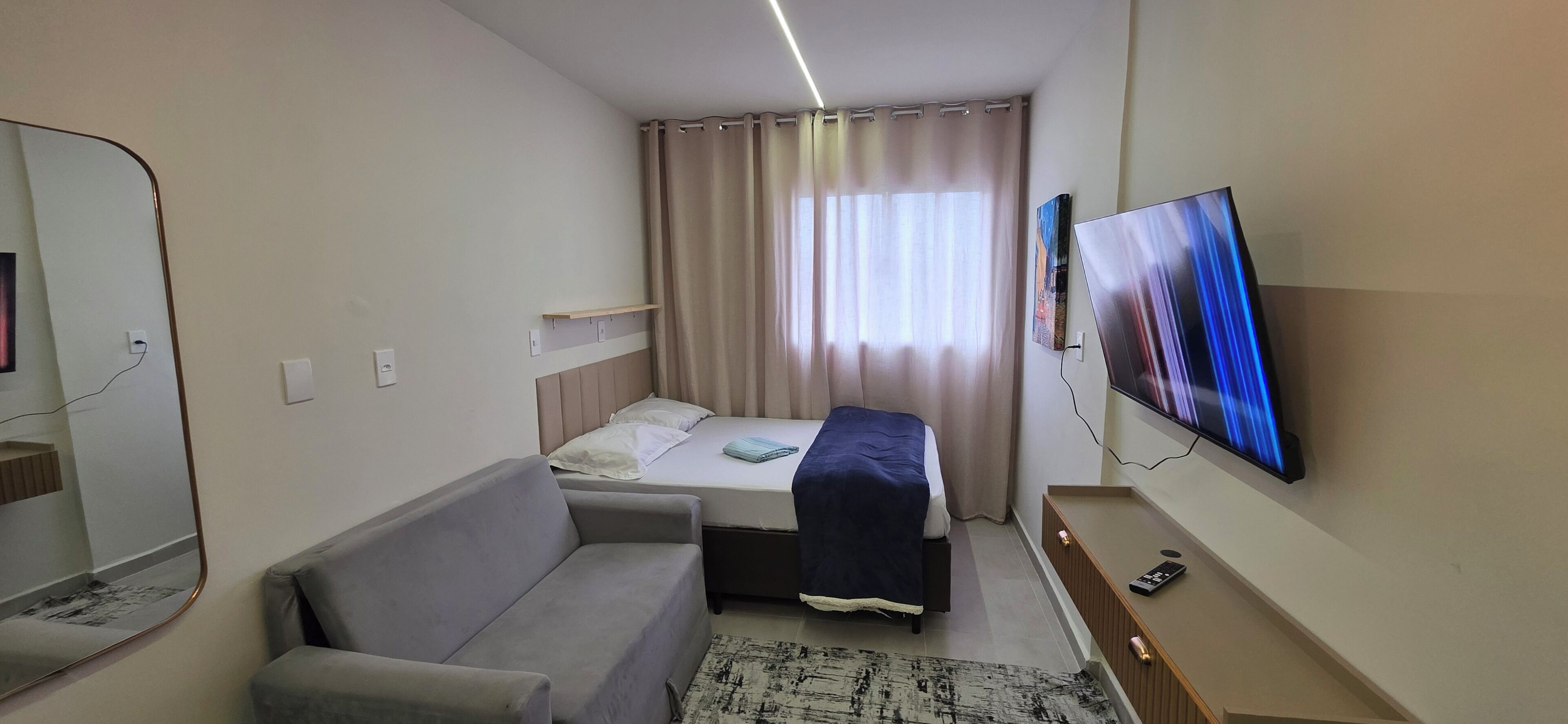 Pieni hotellihuone, jossa on sänky, harmaa sohva, televisio ja peili.