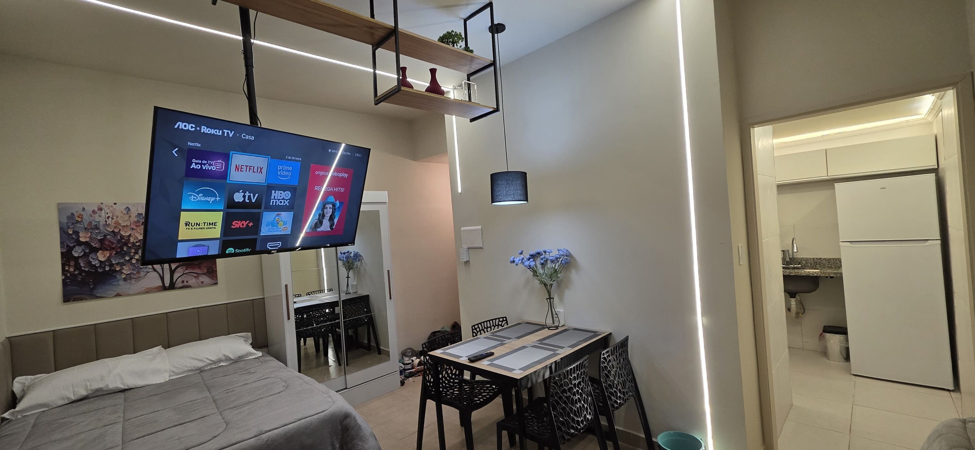 A modern hotel room with a large flat-screen TV displaying streaming services, a bed with a headboard, a dining table with chairs, and a small kitchen area.