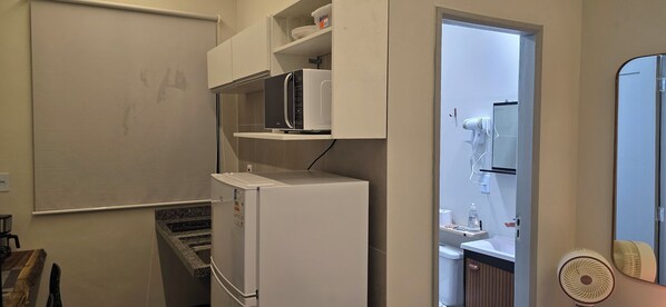 A compact kitchen with a built-in microwave, a white refrigerator, and a small sink. There is a white cabinet above the refrigerator, and a white fan is visible in the corner. - Delta Acomodações (São Paulo)
