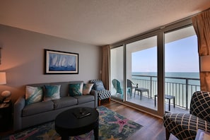 A modern living room with a gray sofa, a round coffee table, and a large window offering a view of the ocean. - Spacious 5-Bedroom Oceanfront Condo in Myrtle Beach (Myrtle Beach)