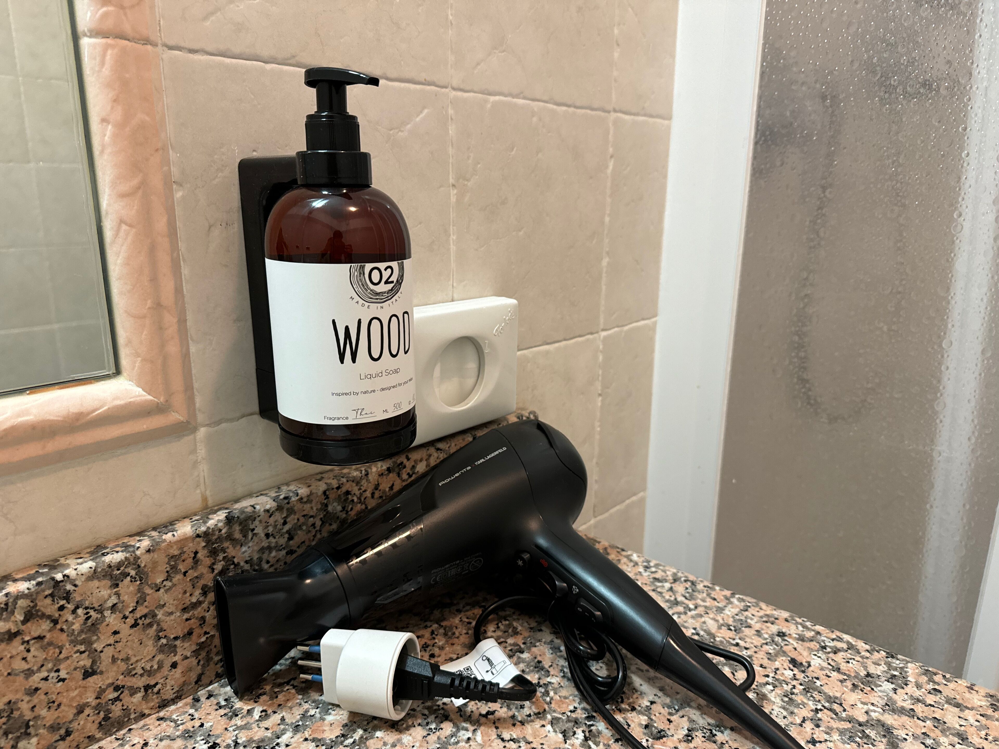 Une salle de bain équipée d’un sèche-cheveux, d’un flacon de savon liquide et d’un distributeur de savon, le tout posé sur un comptoir en granit.