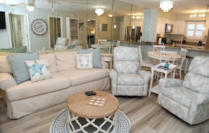 Ein Wohnzimmer mit einem beigefarbenen Sofa, gemusterten Sesseln, einem runden Couchtisch und einer Küche im Hintergrund.