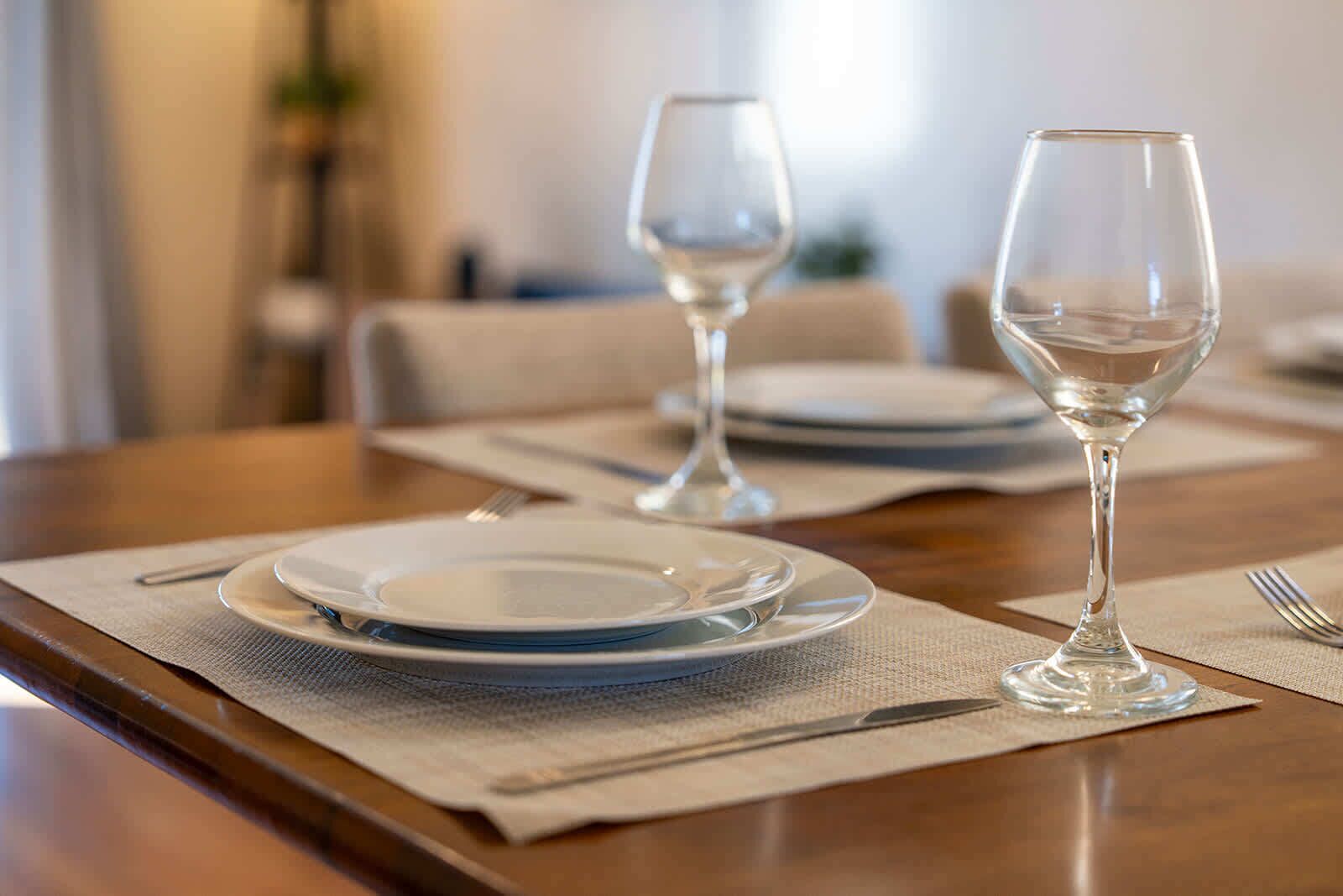 A dining table set with plates, cutlery, and wine glasses, with a placemat and a vase in the background.