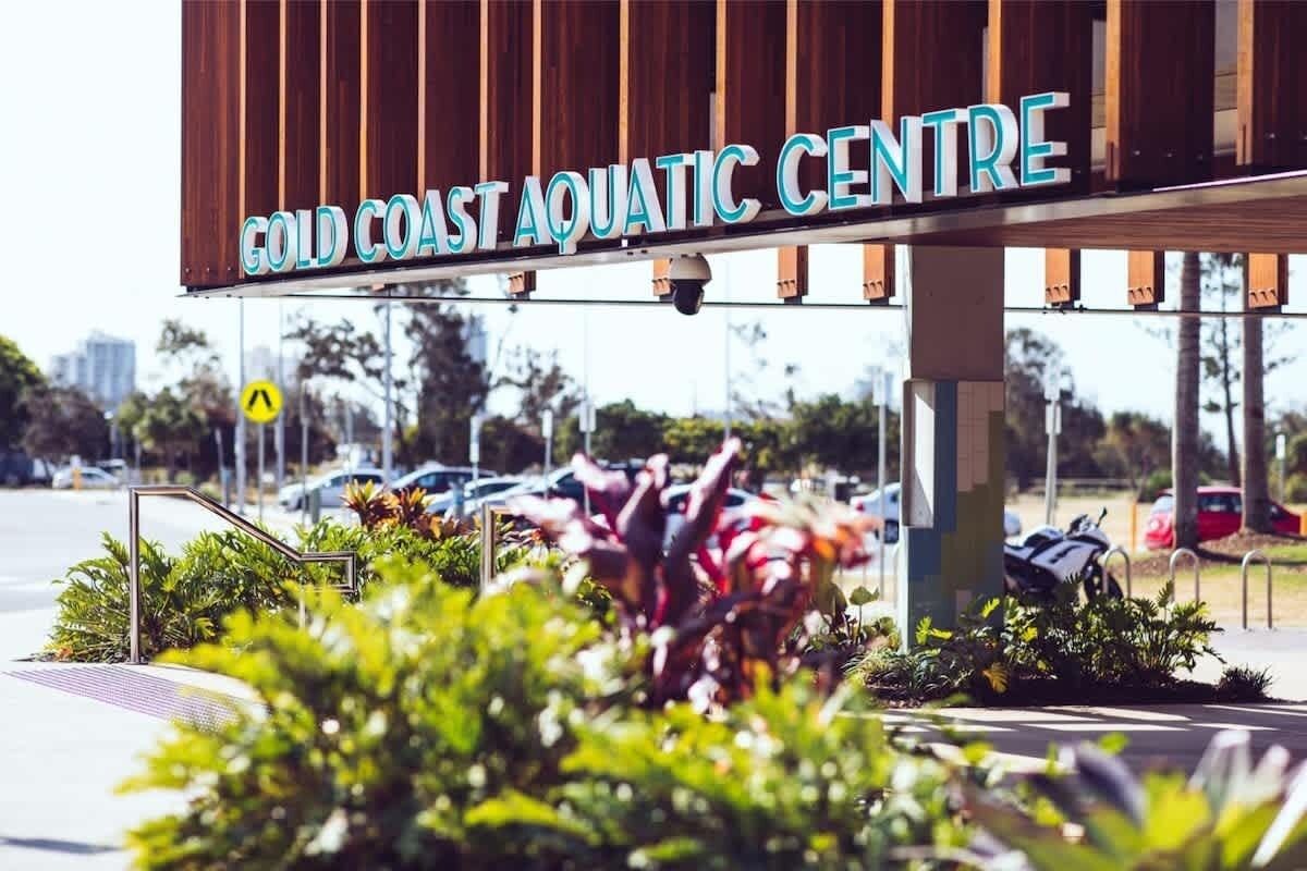 A sign for the Gold Coast Aquatic Centre with a view of the surrounding parking area and greenery.