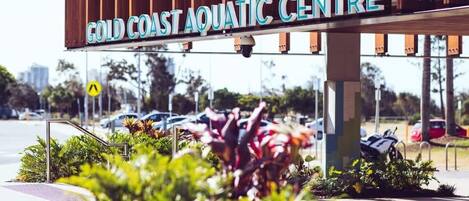 A sign for the Gold Coast Aquatic Centre with a view of the surrounding parking area and greenery.