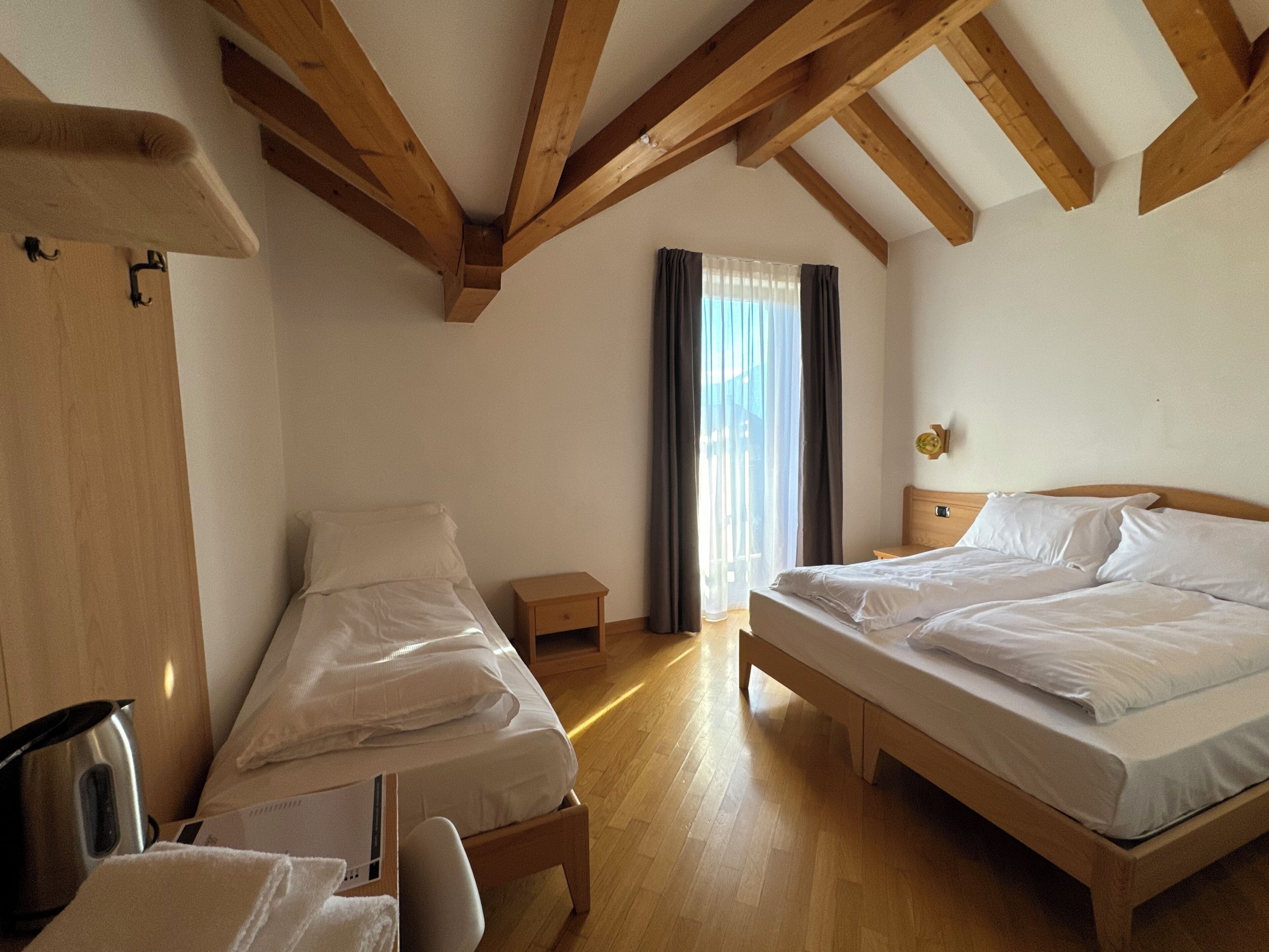A room with two single beds, a small bedside table, a wooden floor, and a window with curtains.