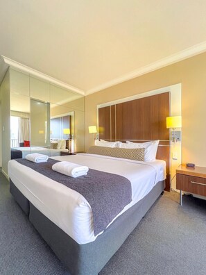 A hotel room with a large bed, a wooden headboard, two bedside lamps, a nightstand, and a large mirror. - Gold Coast Ocean View Apartment with Balcony (Surfers Paradise)