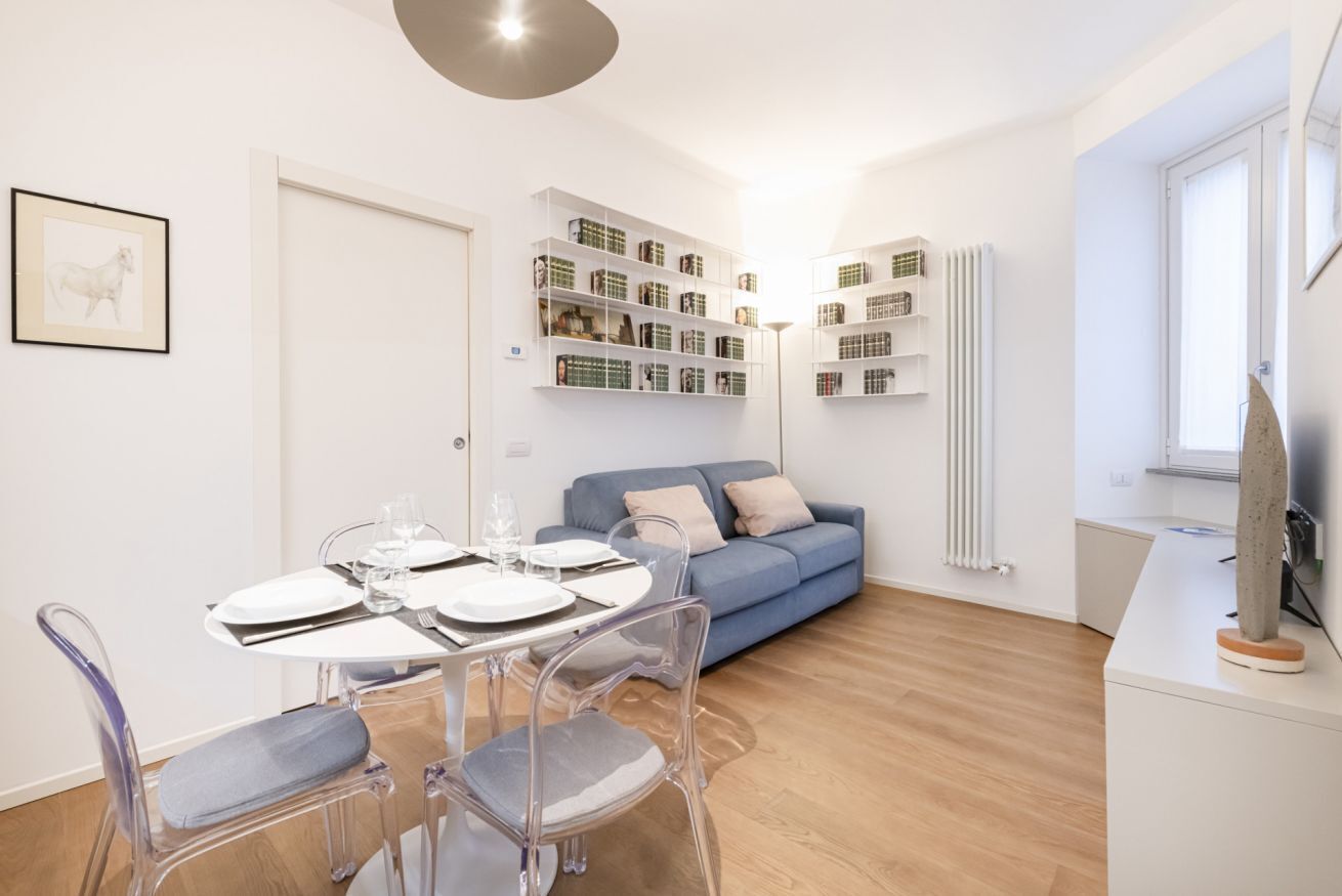 Un soggiorno moderno con un tavolo da pranzo apparecchiato per quattro persone, un divano blu e scaffali a parete con libri e oggetti decorativi.