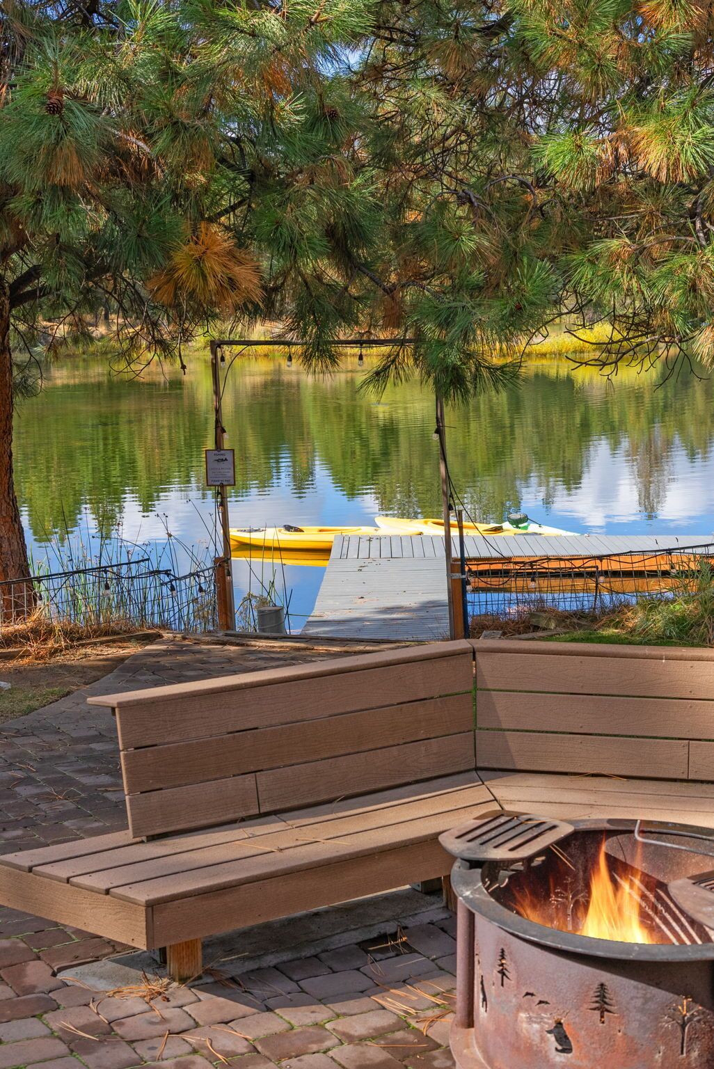 A wooden bench and a fire pit by a calm lake with a dock and a sign.