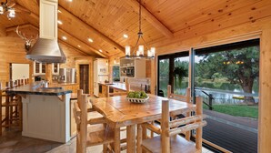 A wooden cabin interior with a dining area, kitchen, and large windows overlooking a lake. - Risa Cabin: Hot Tub, Private Lake (Bend)